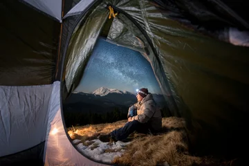 Fotobehang View from inside a tent on the male tourist have a rest in his camping in the mountains at night. Man with a headlamp sitting near campfire under beautiful night sky full of stars and milky way © anatoliy_gleb