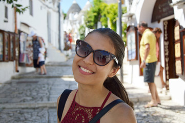 Close up of stylish young woman with sunglasses smiling in italian scenery background. Beauty woman with white perfect smile looking at camera in travel to Alberobello trulli village, Italy, Europe