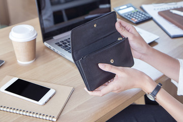 Woman opening her black leather purse to make a purchase online at the office with a cup of coffee, smart phone and laptop on wooden desk.