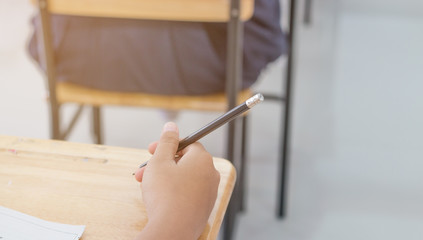 Blurred of Asian students hand holding pen for writing Exams paper sheet or test papers on row wood desk table with student uniform in exam class room, education concept