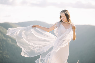 Fototapeta na wymiar Beautiful girl bride in a long dress with curly in the wind boudoir robe, smiling, looking down on the mountains, nature, hair decoration, forest