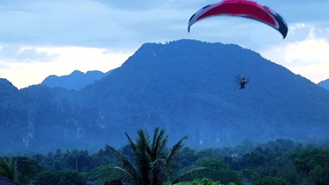 Hang glider hovering over tropical forest. Motorized parachutist flying over the jungle, Vang Vieng, Laos. Recreational activity on holiday.