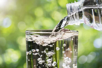 Foto op Plexiglas Close-up van water dat uit de drinkwaterfles in glas stroomt op een wazige groene natuur bokeh achtergrond, gezond drinkwater concept © shark749