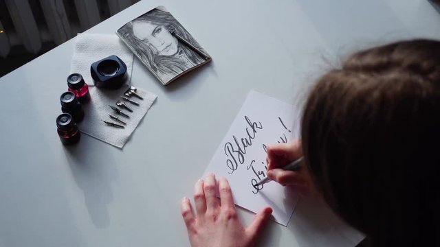 Girl holds brushpen in hands and with beautiful handwriting, wrote black inscription for sales and discounts Black Friday, sitting at white table in art studio. European-looking girl dressed in brown
