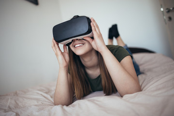 Beautiful young woman lying on the bed and enjoying virtual reality at her home.