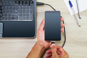 Hands connecting smartphone to laptop by using usb cable on wooden desk with pen and paper, business concept