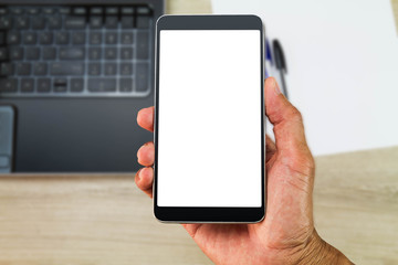 Hand holding white blank screen smartphone above blurred office wooden desk with laptop computer,paper and pens, business concept