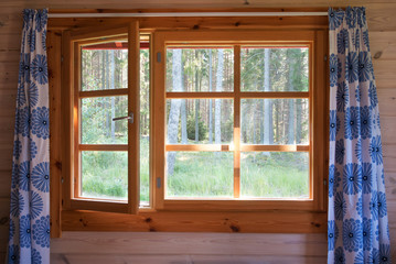 Open window with natural wooden frame and blue flower curtains. Morning forest view. Sunrise in the country