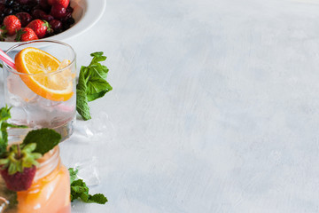 Background with fresh fruit cocktails preparing. Glass of cold drink with citrus slice, mint and ice, berries in bowl ready to use, refreshment and summer concept, close up picture with free space
