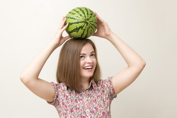 Portrait photo of a smiling pretty girl in pink shirt or blouse in flowery print holding a whole watermelon on her head. White background, warm colors