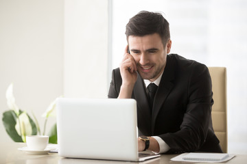 Smiling businessman talking on cellphone sitting at office desk in front of laptop, having successful mobile negotiations with client, manager consulting customer by phone, easy internet banking