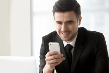 Obraz na płótnie Canvas Attractive smiling businessman looking at smartphone in his hand, texting, sending messages on cellphone, using easy banking app, checking daily calendar or personal organizer, mobile office. Portrait
