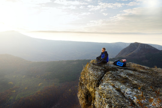 Smiling man hiker with backpack siting and relaxing on the top of the mountain and looking at beautiful yellow autumn landscape sunset over clouds