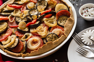Homemade vegetable pie (galette) with grilled eggplants, tomatoes and onion on brown wooden background. Selective focus