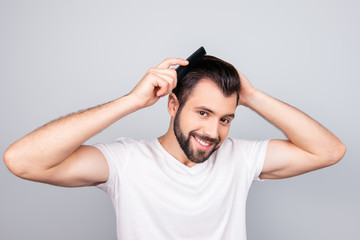 Barber shop concept. Smiling handsome young brunet in white t shirt is standing on the pure background, brushing his hair. His hairdo is stylish and trendy