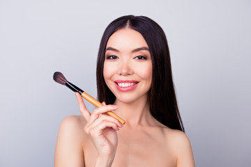 Sensual pretty asian young woman is holding make up brush for powder, smiling, standing on pure background