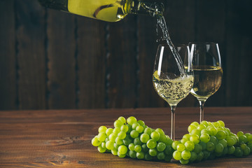 Pouring white wine from the bottle into a glass with a bunch of green grapes against wooden background with free space - obrazy, fototapety, plakaty
