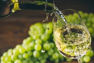 Wall murals Wine Pouring white wine into a glass with a bunch of green grapes against wooden background