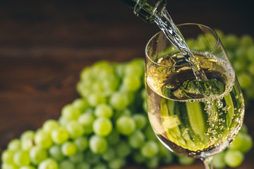 Pouring white wine into a glass with a bunch of green grapes against wooden background - obrazy, fototapety, plakaty