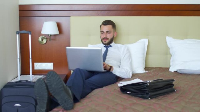 Young businessman in formal wear lying on bed in hotel apartment and using laptop