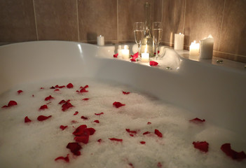 Glasses of alcoholic drink with candles on bathtub filled with foam and rose petals