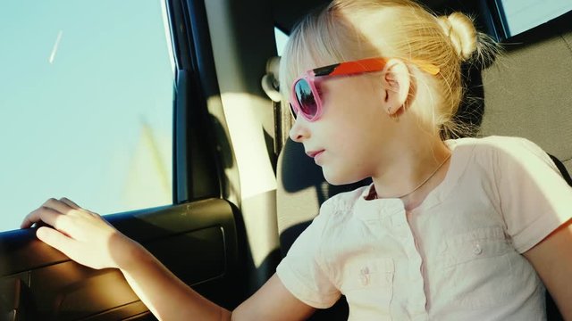 A 6-year-old girl in sun-protective pink glasses is riding in the back seat of the car. It is fastened with a seat belt