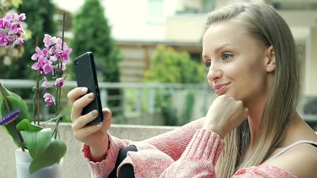 Pretty girl in pink sweater doing selfies on smartphone on terrace, steadycam shot
