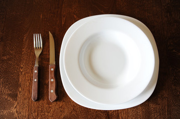 Empty white plate with a fork and knife on a dark wooden brown table