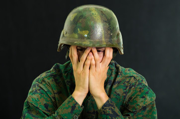 Handsome young soldier wearing uniform suffering from stress, using both hands to cover his face, in a black background