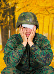 Handsome young soldier wearing uniform suffering from stress, with a white bandage around his head and covering his eye, with both hands in his cheeks, in a blurred yellow background