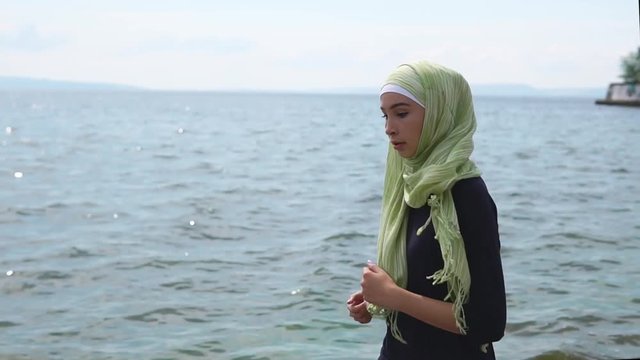 Slow motion steadicam shot of a young Muslim woman having jog training near the sea. She wearing sportswear and green hijab. Healthy lifestyle