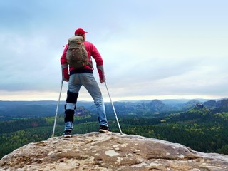 Disabled man with crutches stands on a big rock and looking to mountains at horizon.