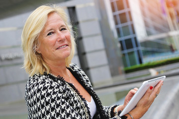 portrait femme senior active et souriante travaillant avec tablette devant centre d'affaires avec buildings 