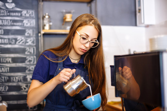 The girl adds milk to the cup with coffee in cafe. Young woman barista working in coffee shop