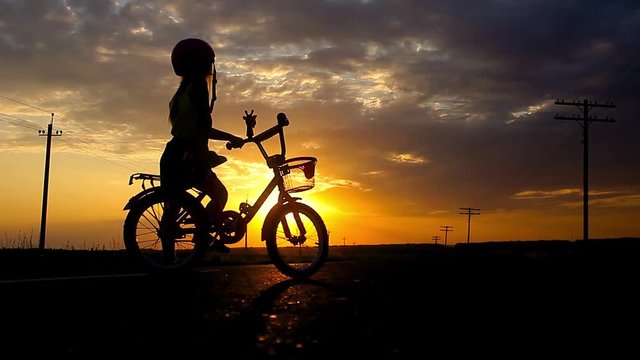 Little girl having fun riding bike at sunset, active family sport, active kids sport, Silhouette a kid at the sunset, Moments of family happiness. Orange-blue sky background.