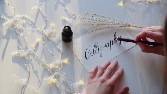 Close-up shot of girl's hands, which displays letters in italic type in calligraphy word, using black ink and fountain pen for calligraphy and writes beautifully italic letters and words, sits at