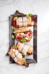 Fresh baguette sandwich bahn-mi styled. Bacon, roasted cheese, tomatoes and lettuce on metallic tray on white marble background. Vertical composition.