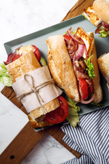 Fresh baguette sandwich bahn-mi styled. Bacon, roasted cheese, tomatoes and lettuce on metallic tray on white marble background.