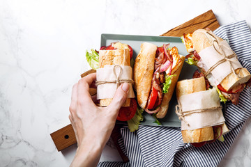 Fresh baguette sandwich bahn-mi styled. Bacon, roasted cheese, tomatoes and lettuce on metallic tray on white marble background. Female hand holding sandwich top view.