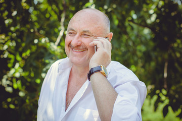 Mature stylish European or American man wear in white shirt at garden, happy senior talk to telephone and hear good news