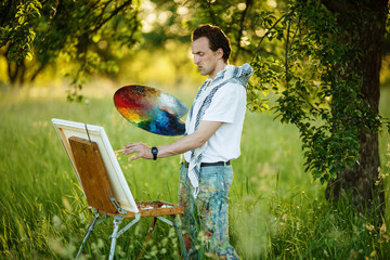 Fototapeta premium Male artist on painting on canvas in outdoor. Young artist painting a landscape. Plein air painting.