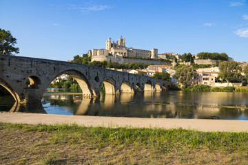 Fototapeta premium Views at sunset of the French city of Beziers, with trees and the old bridge reflected over the river Orb, and the 13th-century Cathedral of Saint Nazaire in the background