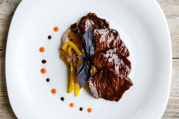 A white plate with slices of beef, stewed yellow pepper and onion. Purple basil on top. Drops of sauce.