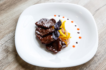 A white plate with slices of beef, stewed yellow pepper and onion. Purple basil on top. Drops of sauce.