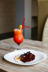 A white plate with slices of beef, stewed yellow pepper and onion. Purple basil on top. Drops of sauce. A colourful red and orange cocktail with a slice of kiwi and two red straws.