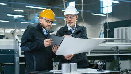 Two engineers discuss a blueprint while checking information on a tablet computer in a factory.
