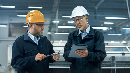 Two engineers in hardhats discuss information on a tablet computer while standing in a factory.
