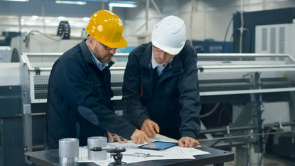 Two engineers discuss a blueprint while checking information on a tablet computer in a factory.