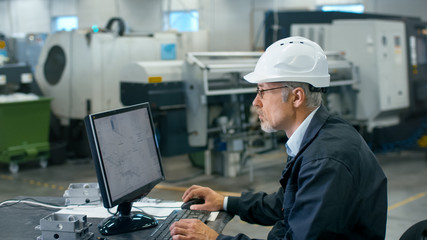 Senior engineer in glasses is working on a desktop computer in a factory.