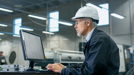 Senior engineer in glasses is working on a desktop computer in a factory.